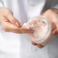 Dłonie w białym kitlu pobierają palcem drobnoziarnisty peeling. W słoiczku widać jego bogatą konsystencję, która może zawierać nawilżającą **urea**.