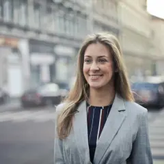 Uśmiechnięta kobieta w marynarce na tle miasta. Czytała pozytywne renee opinie forum i postanowiła wybrać się na spacer.