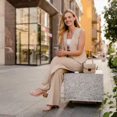 Uśmiechnięta kobieta w beżowym garniturze w stylu smart casual siedzi na ławce, trzymając telefon. Obok niej torebka i słuchawki.