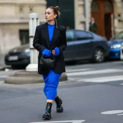 Kobieta w niebieskiej sukience i czarnej marynarce, z czarną torebką. Idealne buty do niebieskiej sukienki? Czarne botki na grubej podeszwie.