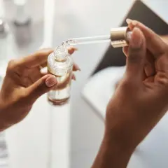 Dłonie trzymają buteleczkę z serum. Pipeta z kroplą produktu, być może zawierającego kwas ferulowy, jest gotowa do aplikacji.