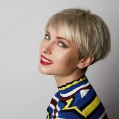 Kobieta z blond pixie bob, w kolorowym swetrze, z czerwonymi ustami, patrzy w stronę aparatu.