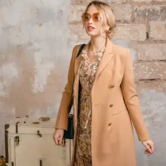 Elegancka kobieta w płaszczu w kolorze camel, sukience w kwiaty i okularach przeciwsłonecznych pozuje przy starej walizce.