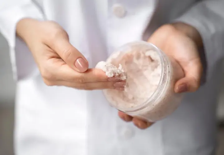 Dłonie w białym kitlu pobierają palcem drobnoziarnisty peeling. W słoiczku widać jego bogatą konsystencję, która może zawierać nawilżającą **urea**.