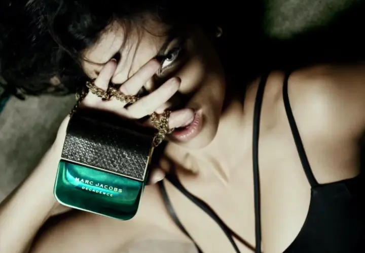 Kobieta zmysłowo trzyma flakon perfum Marc Jacobs Decadence, otoczona złotym łańcuszkiem.