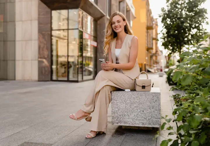 Uśmiechnięta kobieta w beżowym garniturze w stylu smart casual siedzi na ławce, trzymając telefon. Obok niej torebka i słuchawki.