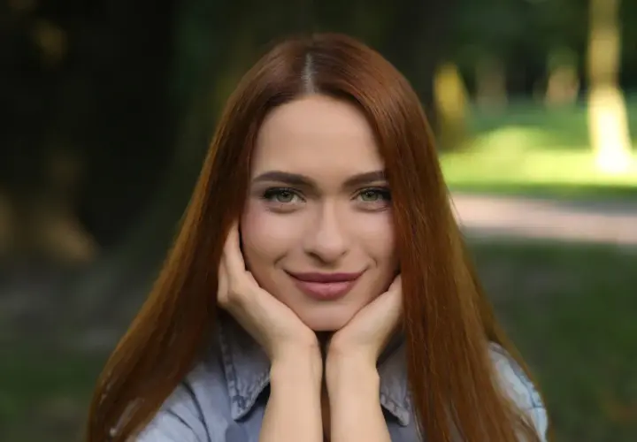 Uśmiechnięta szatynka z rudymi włosami opiera podbródek na dłoniach, patrząc wprost przed siebie.