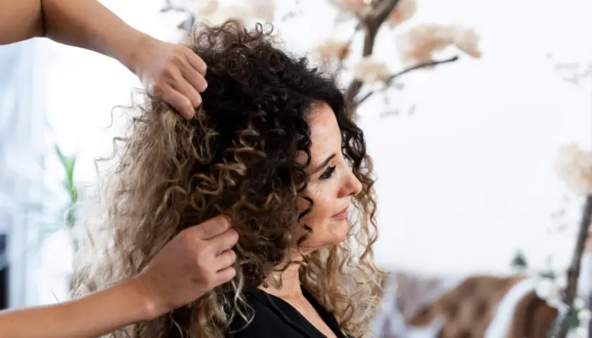 Stylista układa bujne, kręcone włosy kobiety. Jej loki, w odcieniach brązu i blondu, tworzą efektowny bob.