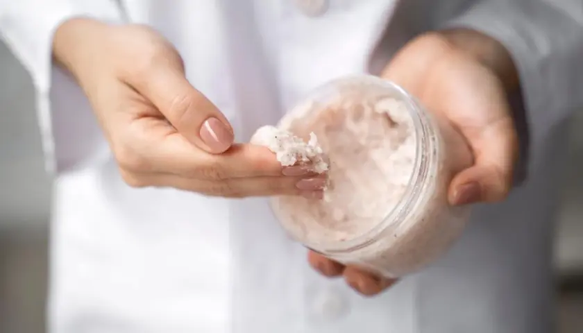 Dłonie w białym kitlu pobierają palcem drobnoziarnisty peeling. W słoiczku widać jego bogatą konsystencję, która może zawierać nawilżającą **urea**.