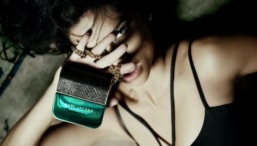Kobieta zmysłowo trzyma flakon perfum Marc Jacobs Decadence, otoczona złotym łańcuszkiem.