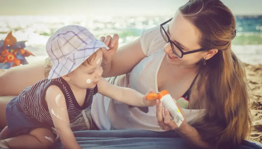 Mama smaruje dziecko kremem z filtrem na plaży. Kolastyna chroni delikatną skórę malucha przed słońcem.