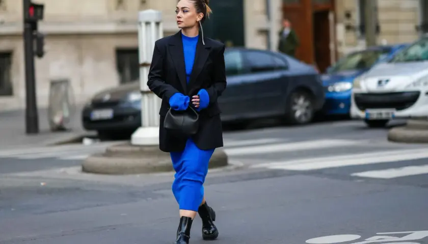 Kobieta w niebieskiej sukience i czarnej marynarce, z czarną torebką. Idealne buty do niebieskiej sukienki? Czarne botki na grubej podeszwie.
