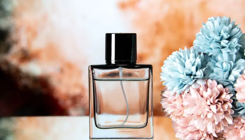 Elegancka butelka perfum z czarną nakrętką, obok bukietu błękitnych i różowych kwiatów. Klik i pachniesz opinie.