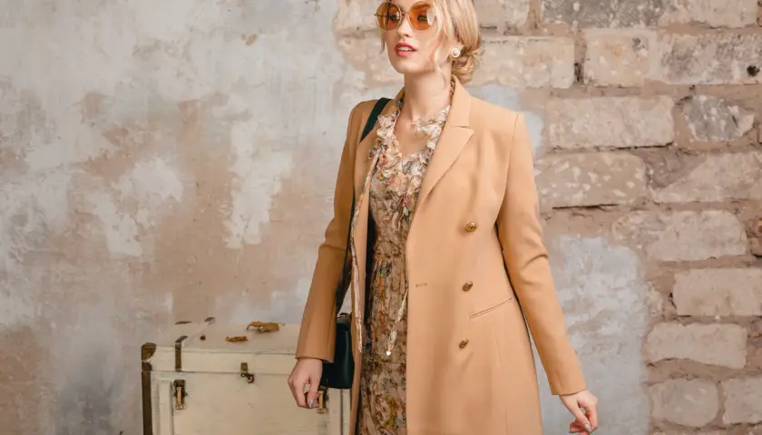 Elegancka kobieta w płaszczu w kolorze camel, sukience w kwiaty i okularach przeciwsłonecznych pozuje przy starej walizce.