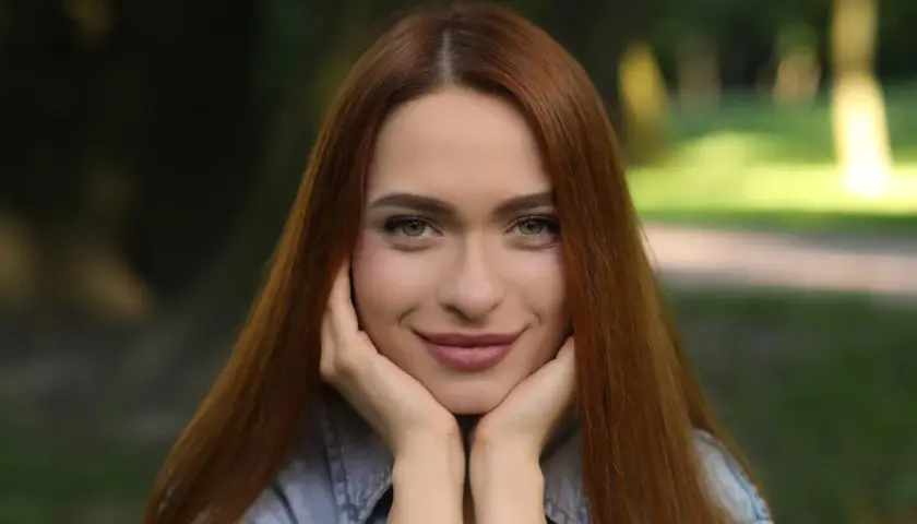 Uśmiechnięta szatynka z rudymi włosami opiera podbródek na dłoniach, patrząc wprost przed siebie.