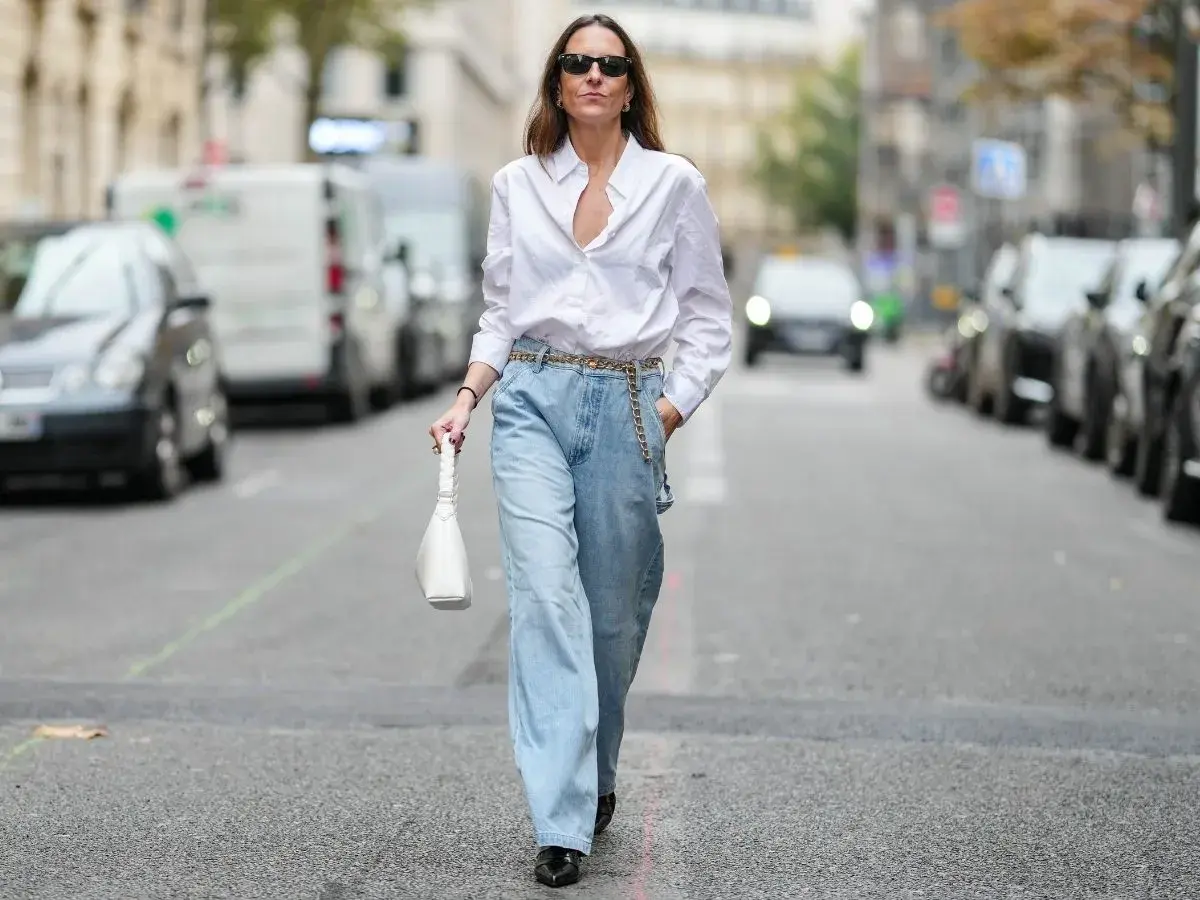 Kobieta w luźnych, jasnych jeansach typu mom fit i białej koszuli, z białą torebką. Stylizacja dla każdej kobiety ceniącej wygodę i modny look.