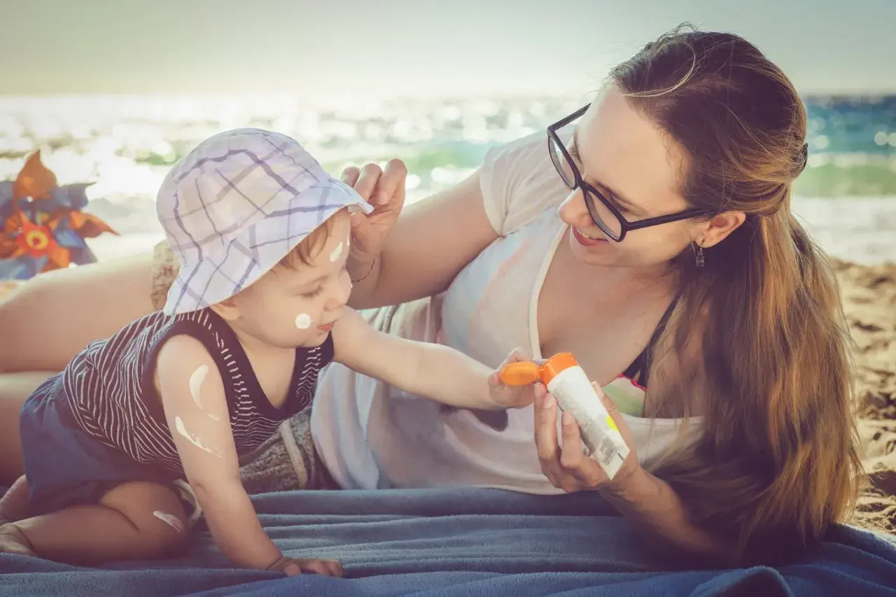 Mama smaruje dziecko kremem z filtrem na plaży. Kolastyna chroni delikatną skórę malucha przed słońcem.