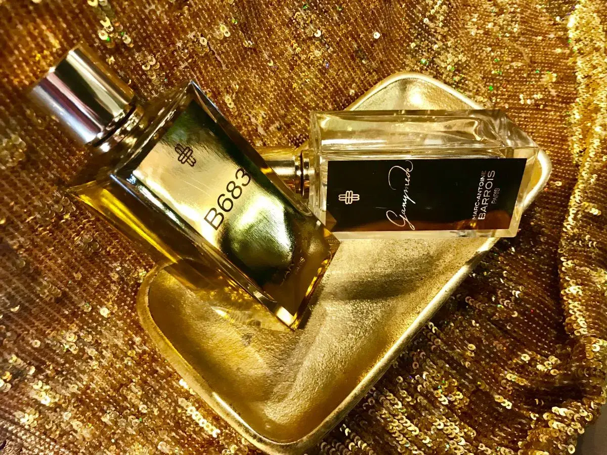 Dwa flakony perfum Marc Antoine Barrois, jeden z napisem B683, drugi z Ganymede, na złotej, cekinowej tkaninie.
