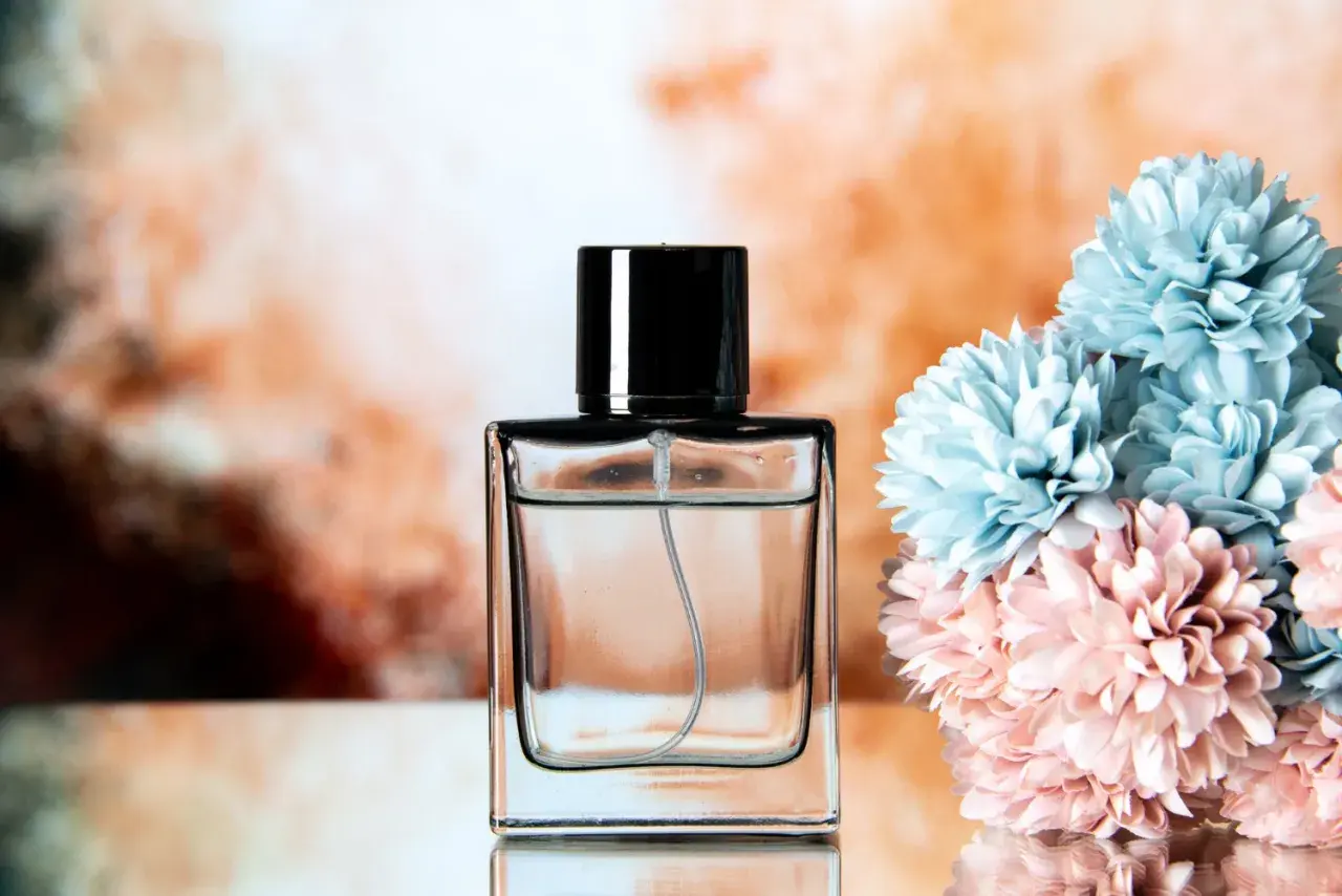 Elegancka butelka perfum z czarną nakrętką, obok bukietu błękitnych i różowych kwiatów. Klik i pachniesz opinie.