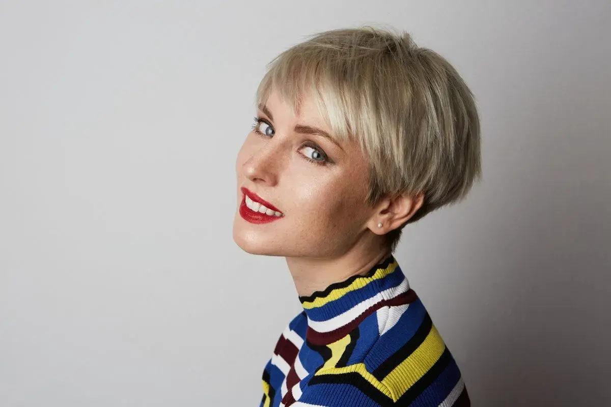 Kobieta z blond pixie bob, w kolorowym swetrze, z czerwonymi ustami, patrzy w stronę aparatu.