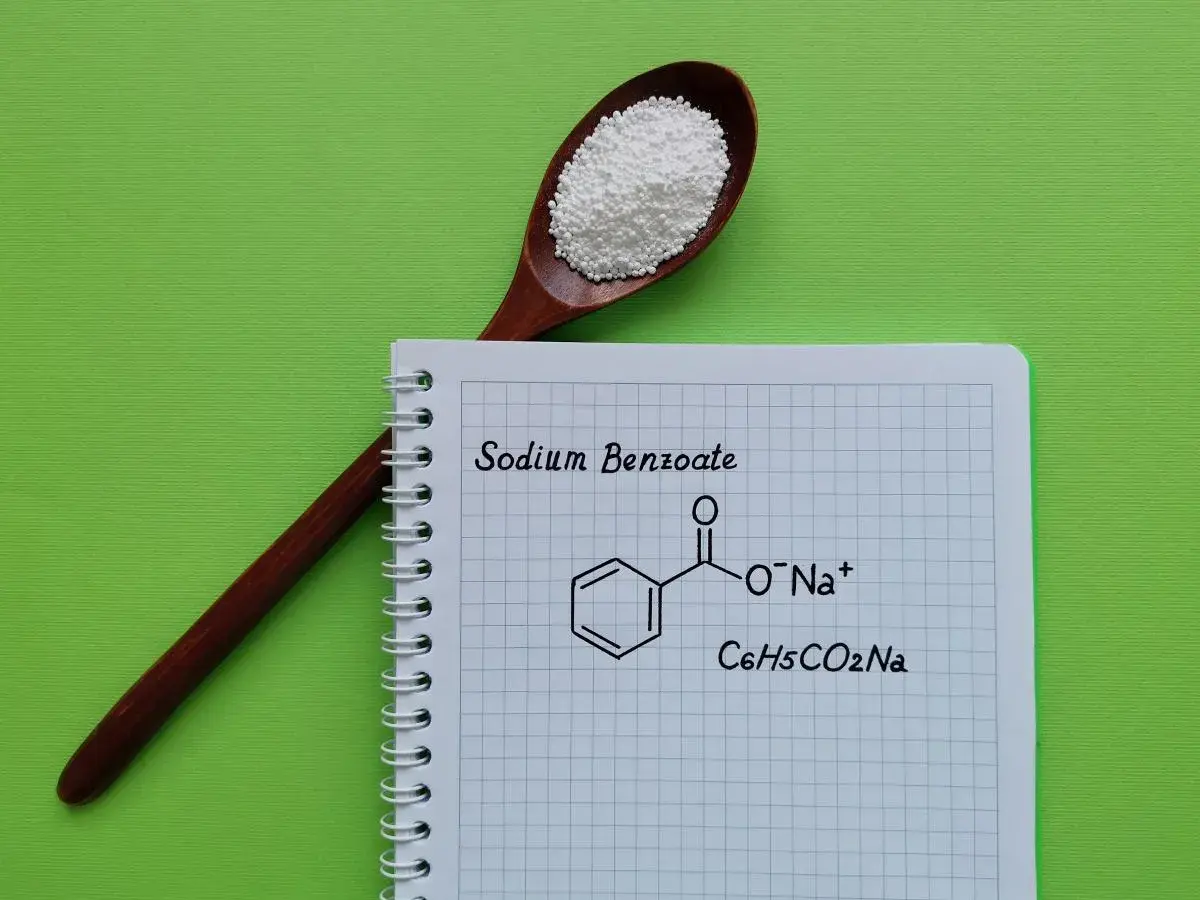 Drewniana łyżka z białym proszkiem, obok notatnik z napisem "Sodium Benzoate" i jego wzorem chemicznym.