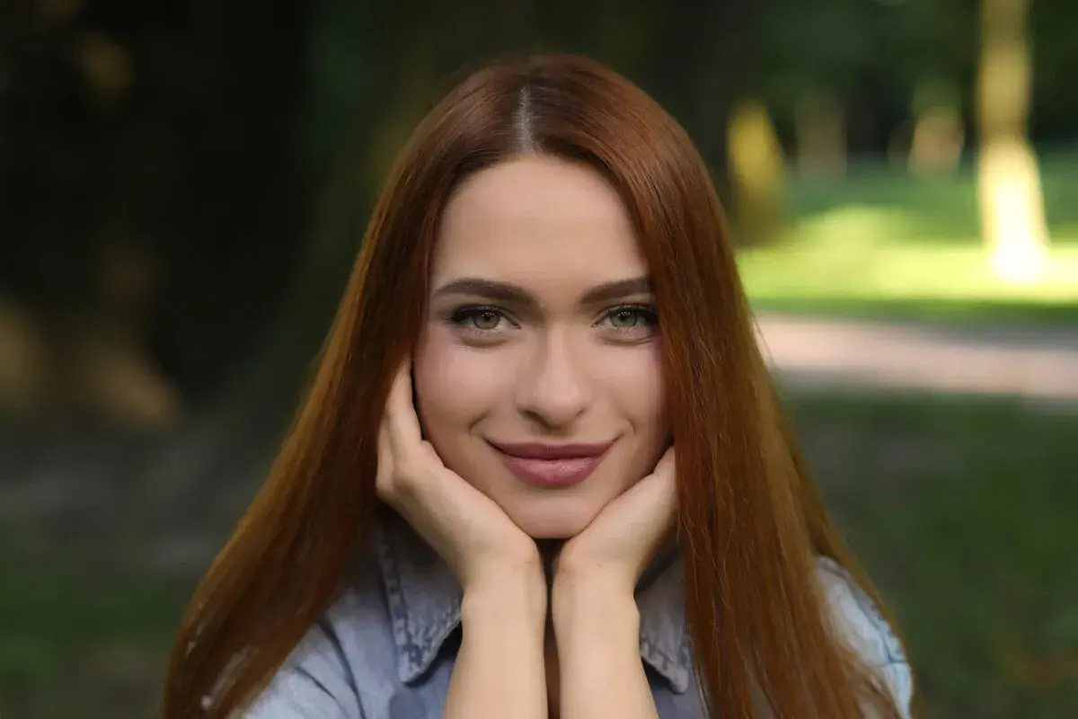 Uśmiechnięta szatynka z rudymi włosami opiera podbródek na dłoniach, patrząc wprost przed siebie.
