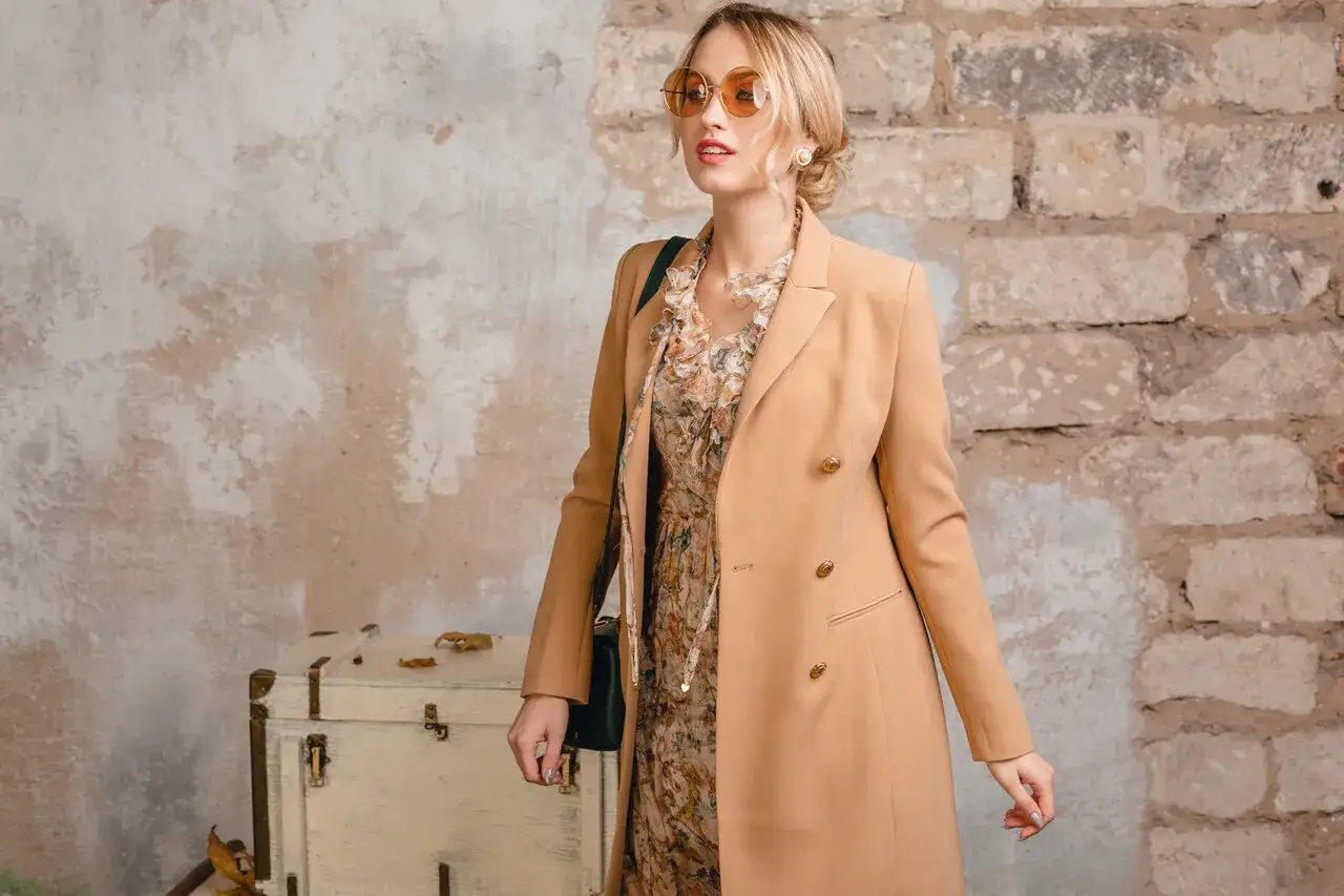 Elegancka kobieta w płaszczu w kolorze camel, sukience w kwiaty i okularach przeciwsłonecznych pozuje przy starej walizce.