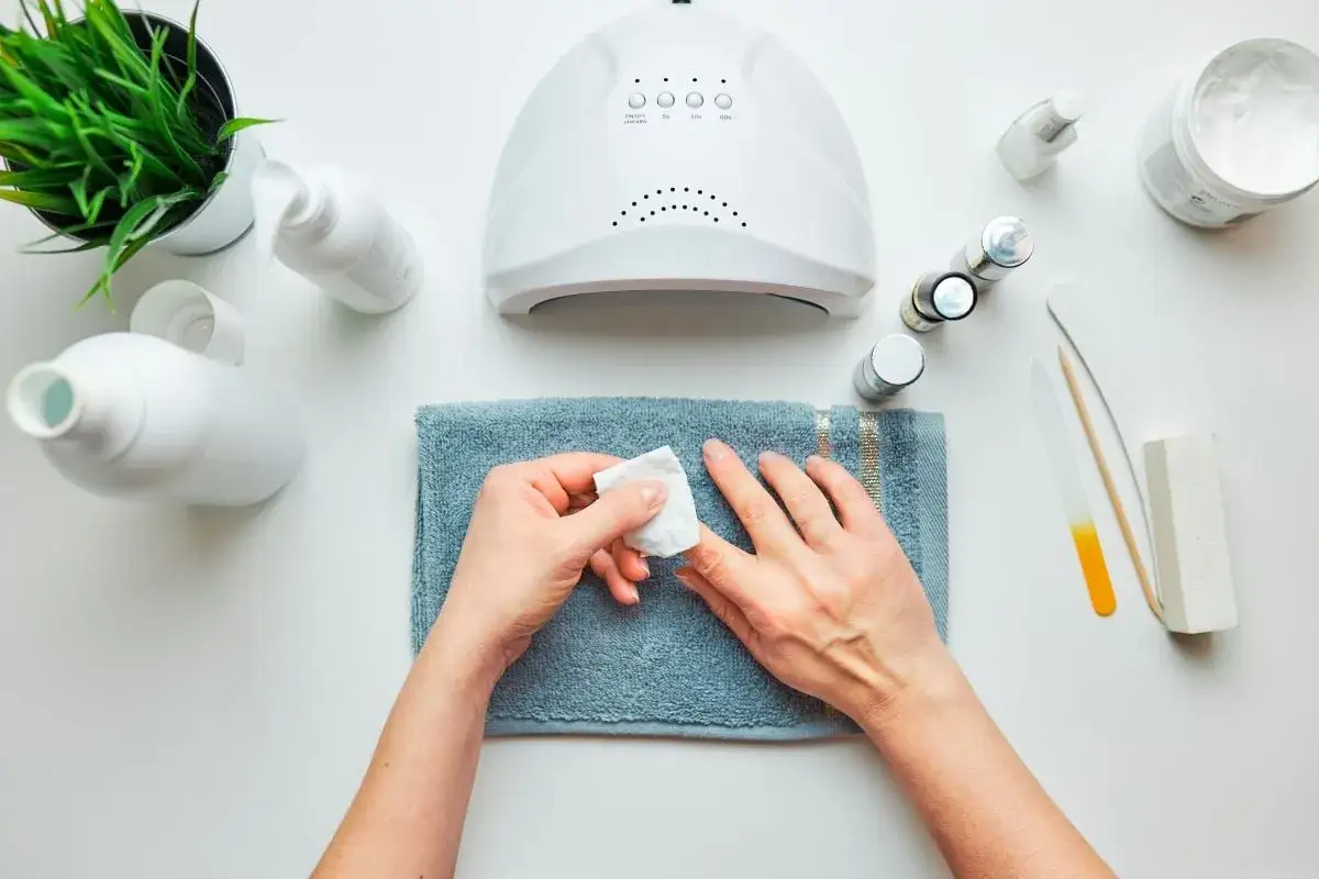 Przygotowanie do manicure hybrydowego: dłonie na ręczniku, lampa UV, lakiery i akcesoria do paznokci.